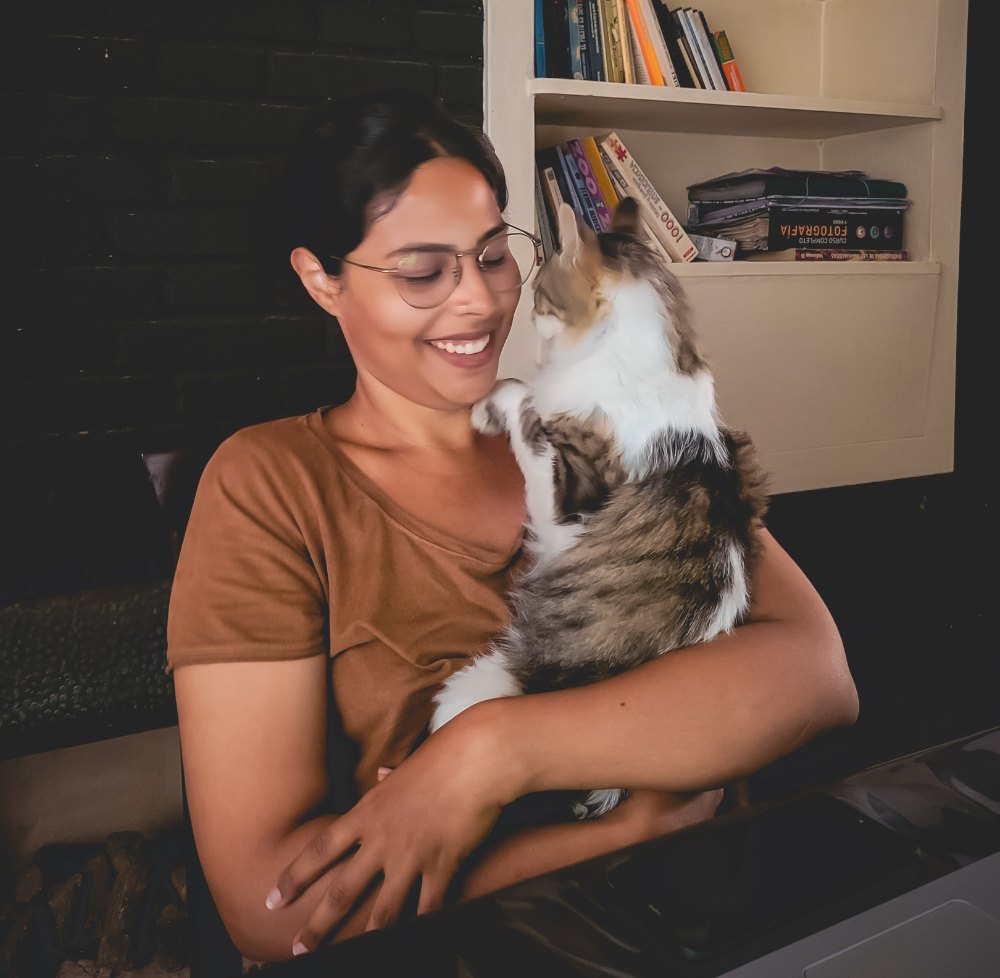 Isabella sorrindo, com um gato de pelo longo no colo, diante de uma estante de livros.